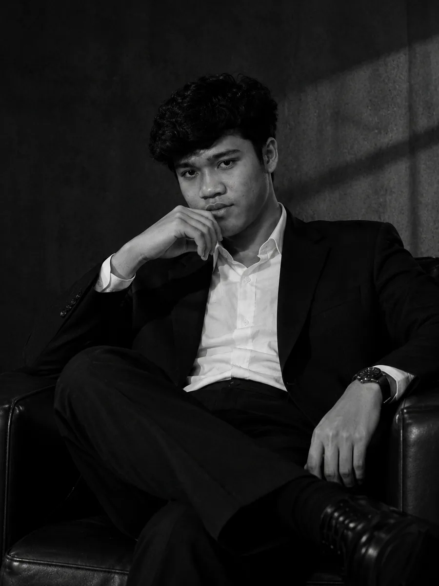Foto Ach Muhlis (Austin Official) duduk santai di kursi kulit, berjas hitam dan kemeja putih